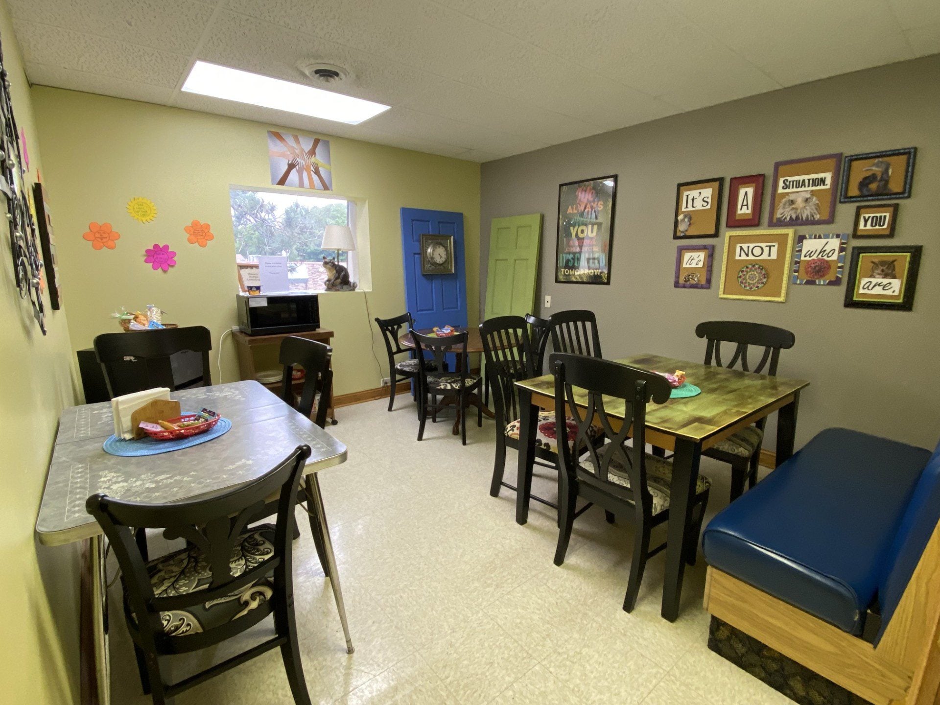 Cozy break room with tables, chairs, a blue door, framed motivational quotes on the wall, flower cutouts, and a window with an outdoor view. Food and drinks are set on the tables, creating a welcoming space designed to offer support for youth in Central Minnesota.