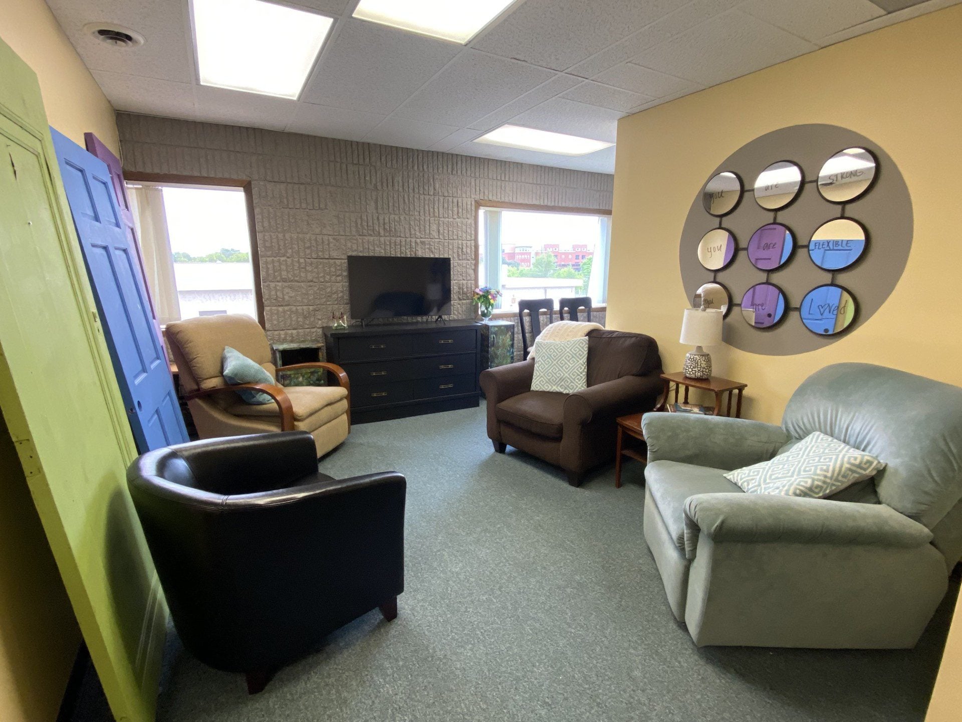 A cozy office lounge with armchairs, a TV on a dresser, decorative mirrors on a yellow wall, and sunlight streaming through windows in the background—an inviting space dedicated to providing support for youth in Central Minnesota.