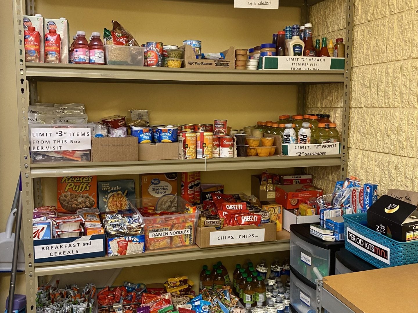 Shelves stocked with canned foods, snacks, drinks, fruit cups, ramen, cereal, and hygiene items provide vital support for youth in Central Minnesota. Signs limit the number of items people can take. Boxes are labeled by food type, and some drinks are in bottles.