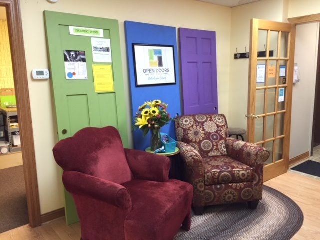 Two colorful armchairs sit on a round rug in front of three painted doors on a wall. A side table holds a vase of sunflowers. The space looks cozy and inviting, with informational flyers about Support for Youth in Central Minnesota displayed on the green door.