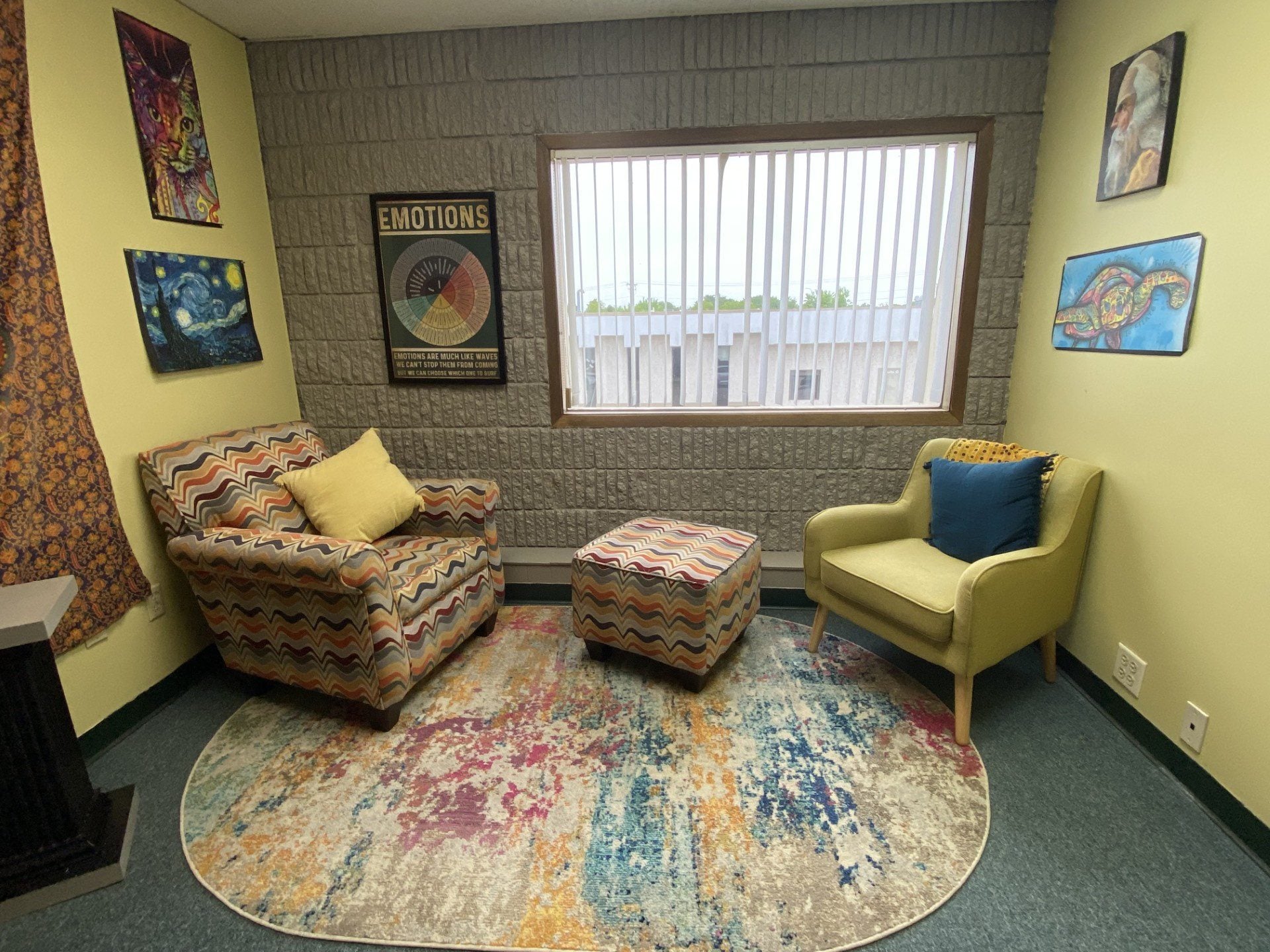 Cozy therapy room with two armchairs, a patterned ottoman, and a colorful rug. Artworks and an emotions chart hang on pale yellow walls, reflecting the supportive environment offered for youth in Central Minnesota. A large window with vertical blinds lets in natural light.
