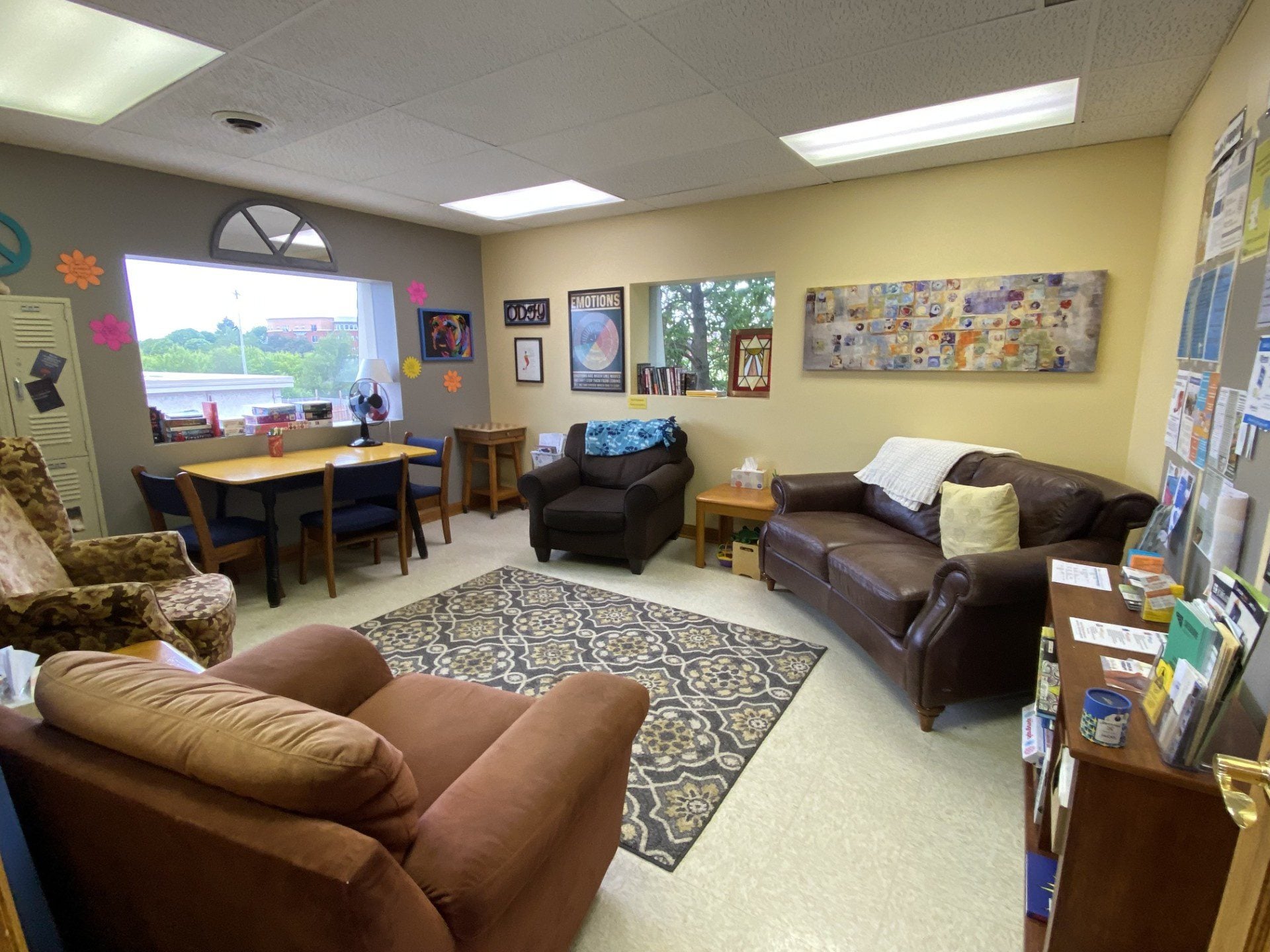 A cozy office with armchairs, a brown leather sofa, a small table, bookshelf, and a rug. Art and flyers decorate the walls; a window lets in natural light. Pamphlets—highlighting support for youth in Central Minnesota—are displayed on a shelf on the right.