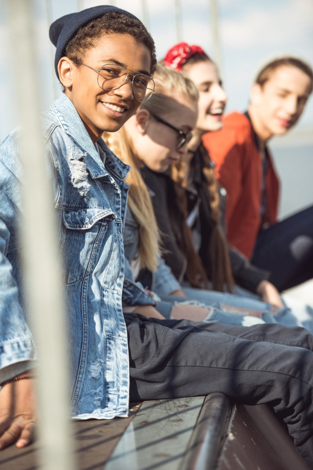 Smiling teenage boy wearing glasses and a denim jacket sits with three friends outdoors, all relaxed and enjoying the sunny weather—a perfect example of support for youth in Central Minnesota, where community and friendship thrive.