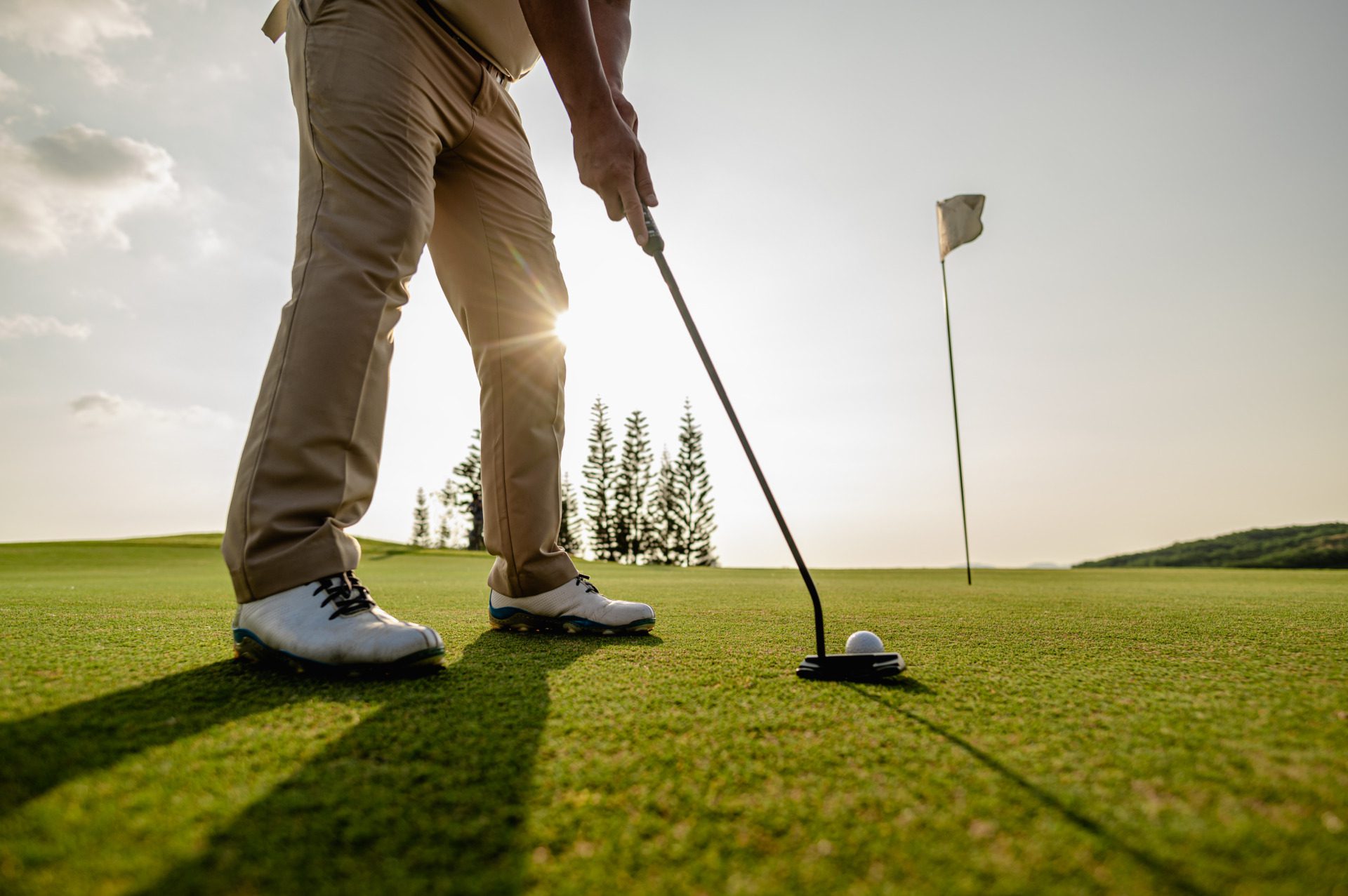 A golfer in beige pants lines up a putt on a green, with the sun low behind him and a flagstick in the distance during the Noelle M. Anderson Memorial Golf Tournament.