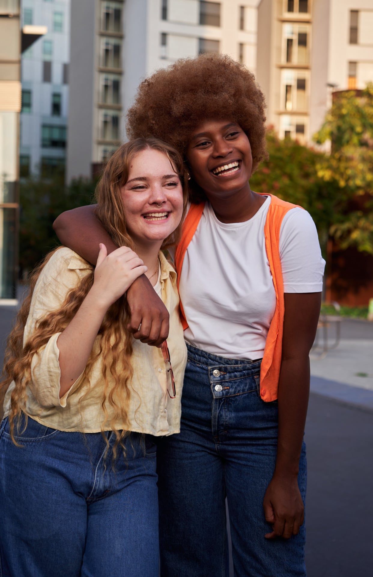 Two young women standing outside, smiling and hugging each other—one in a yellow shirt, the other in a white shirt with an orange vest—embody the spirit of support for youth in Central Minnesota. Urban buildings and greenery provide a vibrant backdrop to their joyful connection.