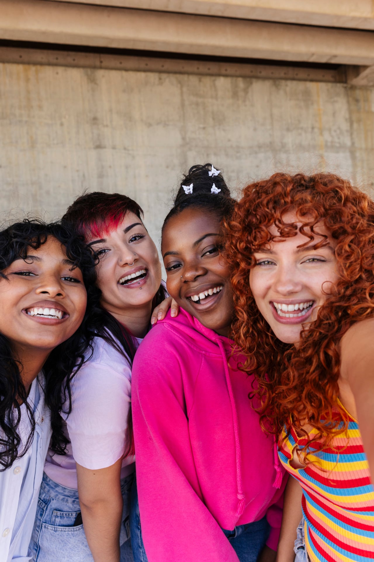 Four young women smile and pose closely together for a selfie, standing outdoors against a concrete wall, embodying the spirit of support for youth in Central Minnesota.